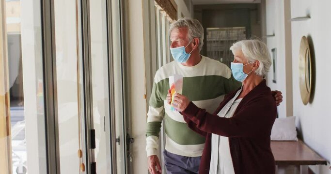 Senior caucasian couple wearing face masks holding rainbow painting against the window at home