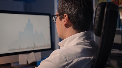Home trader analysing trading charts on stock market on computer screen at home. Pan shot side view of man in eyeglasses looking at the screen with stock graphs