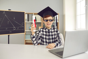 Happy child in academic cap and funny glasses sitting at desk and holding graduate certificate