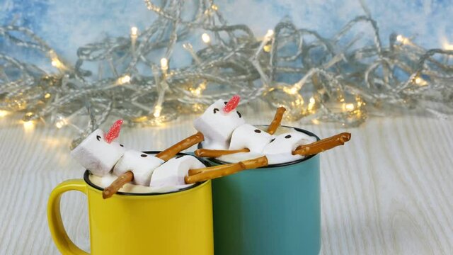 Two sweet snowmen in yellow and blue cups on light background with flashing garland
