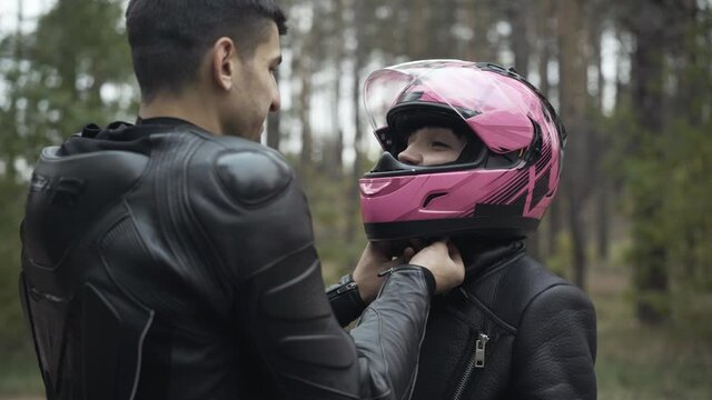 Portrait of young Caucasian girlfriend smiling to boyfriend as Middle Eastern man fastening her pink motorcycle helmet. Loving interracial happy couple of motorbikers preparing for a ride outdoors.