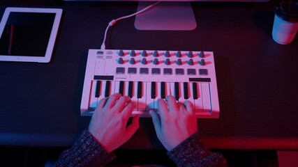 Closeup of hands composing music in night using midi controller. Top view of person playing music with electronic keyboard, midi keys on the table with neon lights © diproduction