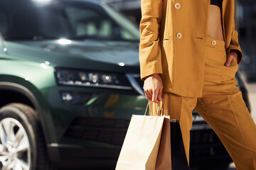 With paper bag in hand. Young fashionable woman in burgundy colored coat at daytime with her car