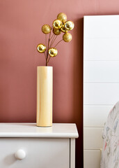 Branch with golden christmas balls in a vase on a nightstand in front of a terracotta painted wall.