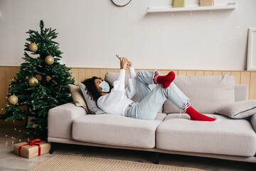 Black individuals concept. Young african american wearing protective mask, typing a message while lying on her sofa in Christmas-decorated room.
