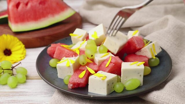 Vegetarian salad with watermelon, feta cheese, and grapes stir with fork on blue ceramic plate on white wooden background and linen textile. Side view, close up, selective focus.