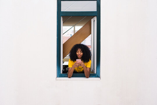 Cheerful ethnic woman using phone while looking out window