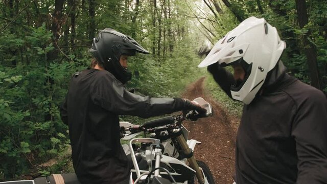 Handheld tracking of cheerful male motorcyclists in helmets high-fiving each other in forest with lush greenery