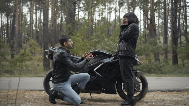 Wide shot of loving confident Middle Eastern man proposing to Caucasian woman outdoors. Handsome biker fiancee putting engagement ring on finger of bride and hugging partner. Love and romance.