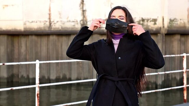 Eine sch&ouml;ne Frau mit langen dunklen Haaren in einem schwarzen Mantel versucht eine schwarze Maske aufzusetzen. Die Maske h&auml;lt aber nicht und f&auml;llt ab.  Die Frau lacht.