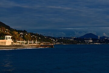 Fototapeta premium Seascape with a jutting cape with a rotunda in the rays of sunset against the background of mountains and the city
