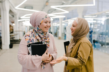 Happy Muslim female colleagues with copybooks chatting in hallway