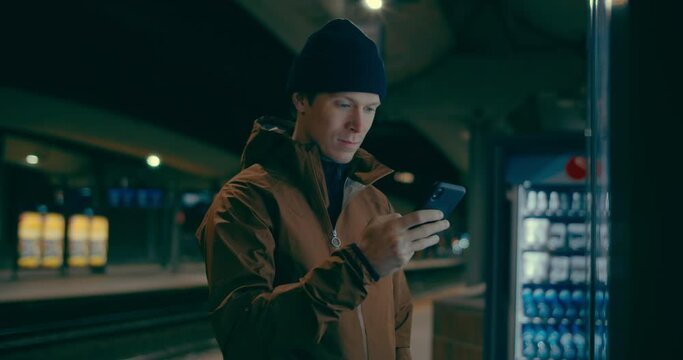 Handsome young millennial man check train schedule on smartphone. Man on subway or train platform at night. Screen lit face, texting and chatting on social media. Big city life, daily commute