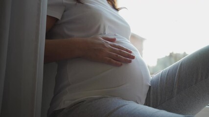 Beautiful pregnant woman sitting by window at home, brunette lady with pretty face touching her belly, pregnancy and motherhood concept. - Powered by Adobe