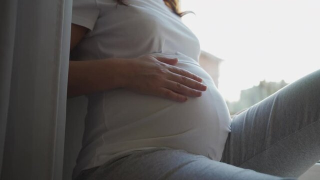 Beautiful pregnant woman sitting by window at home, brunette lady with pretty face touching her belly, pregnancy and motherhood concept.