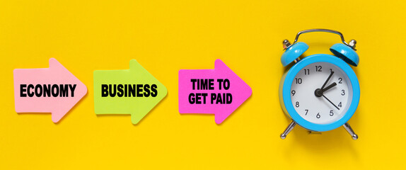 On a yellow background, a blue alarm clock, and paper arrows. On the pink arrow it says - Economy, on the yellow - Business and on the red - TIME TO GET PAID