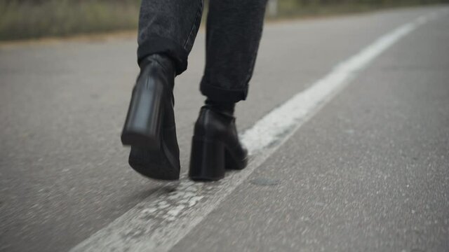 Female feet in jeans and black leather high-heels walking along white line road marking. Unrecognizable young woman strolling on empty highway on cloudy spring or autumn day.