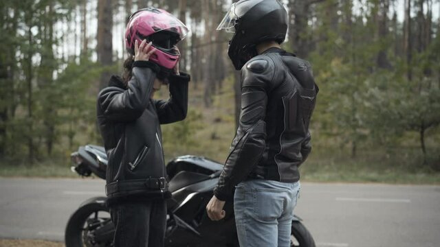 Angry couple of biker arguing outdoors. Irritated young woman taking off pink helmet, giving it to stressed man, and leaving. Conflict of interracial boyfriend and girlfriend on empty roadside.
