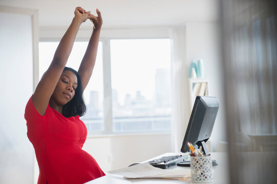 Black pregnant businesswoman stretching arms at desk in office