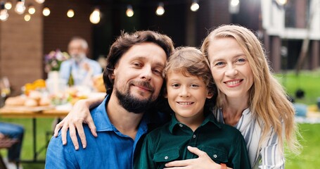 Portrait of Caucasian father and mother hugging small cute son and smiling in yard. Handsome man and beautiful woman embracing little boy. Outdoors. Family dinner. Happiness. Kid with dad and mom.