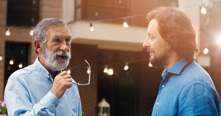 Handsome Caucasian two men, old and young, standing at back yard of house and talking. Family dinner outdoor on background. Senior father with adult son having talk and conversation. Generations.