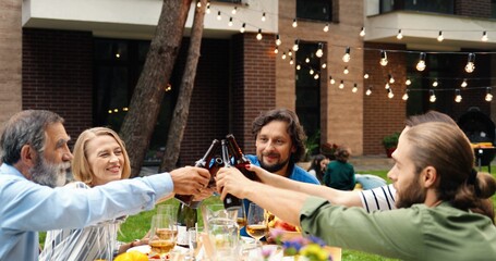 Smiled cheerful Caucasian family laughing, talking and toasting at barbecue outdoor on weekend day. Happy people of different ages saying toasts and cheering with beer bottles. Drinks in hands.