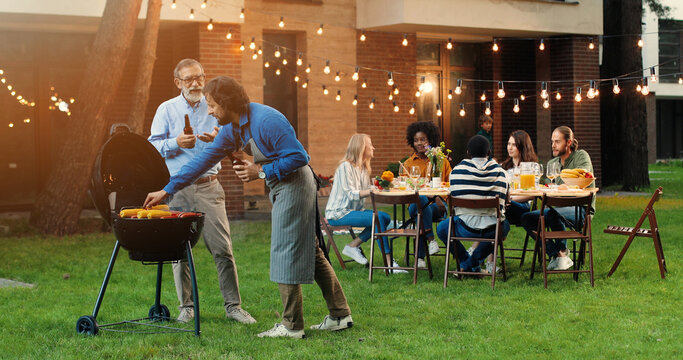 Old gray-haired father and middle-aged son standing outdoor at barbecue on back yard, drinking beer and talking. Caucasian adult man with retired senior dad having nice talk, cooking food on fresh air