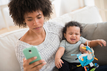 Mother holding crying baby son while texting on cell phone