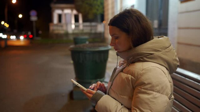 Attractive adult woman in warm coat holding cell phone in the street at cold night, brunette lady typing on mobile phone screen on an empty street. Pavlovsk Park in autumn, St. Petersburg, Russia.