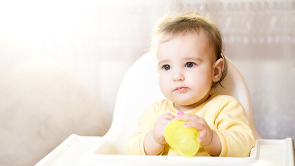 Little baby sitting in high chair and eating food indoors.. baby's first feeding. 6 month old toddler with nibbler in hand.