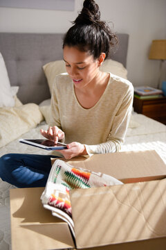 Hispanic woman sitting on bed near box using digital tablet