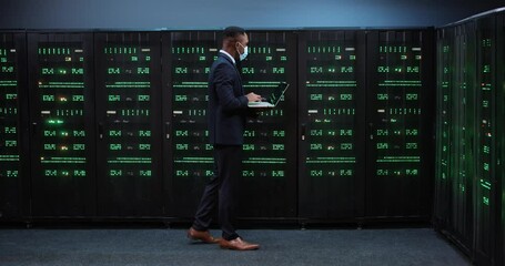 African American server technician specialist in mask typing and browsing on laptop running diagnostics on servers. Man working on data or cyber security in datacenter. Technologies. Hardware concept - Powered by Adobe