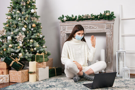 Young woman waving while having a video call with family via computer at home, surrouded by Christmas decorations. New Year theme during covid time. Social distancing during quarantine.