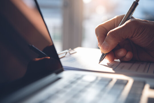 Business man hand with a pen writing on business document with laptop computer on table in office