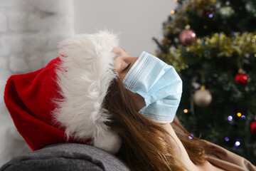Close up image of woman wearing Santa hat and protective face mask relaxing on sofa next to the Christmas tree. Winter holidays during Corona virus outbreak, self-quarantine concept.