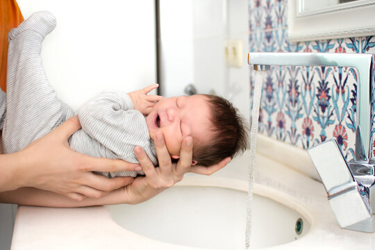 Portrait of adorable cute baby taking his first bath and crying. Water pouring on baby head. First bathing of a newborn baby in a bathtub. First days of life. One week old baby lifestyle photo.
