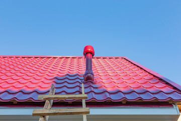 The roof of the house is made of metal tiles against the background of the blue summer sky, there is a wooden staircase and the ventilation outlet is visible.