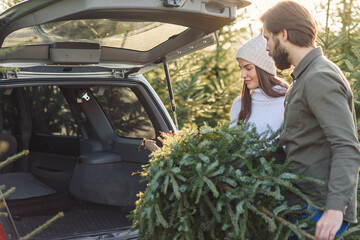 Lovely cheerful young couple putting into car trunk beautiful fir tree at Christmas tree plantings,...