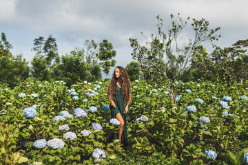 Young beautiful woman in green dress enjoying blooming blue hydrangeas flowers in garden. Gardening and florist concept. Plantation of flowers. Beauty in nature.