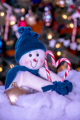 Cute stuffed snowman and candy cane heart, sits in fluffy fake snow by a festive lit Christmas tree
