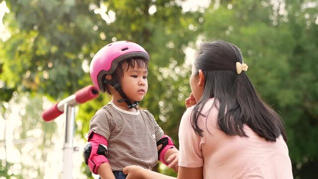 Young mother helping her daughter wears pink helmet before learning to ride scooter in summer park. Active outdoor sport for kids.