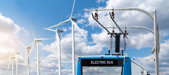Electric bus at a stop is charged by pantograph on the background of wind turbines	
