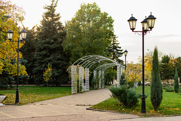 Walkway in the city's public Park. Beautiful white metal arch, lots of greenery, trees and conifers around, lawn grass along the edge, benches and lanterns. The road is tiled