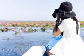 Asian woman wearing a white dress, wearing a hat and sitting on a blue boat. Taking pictures at Lake Of Pink Water Lilies, Nong Han, Kumphawapi, Udon Thani, Thailand.