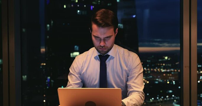 High-ranking official employee work until late in office room sit at workplace desk using laptop prepare report, develop project, solve business issues make urgent task, night city view through window