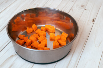 Metal pet bowl with cut carrot inside. First step of natural organic daily meal ration preparation, BARF diet for dogs concept. Copy space, white wooden table background.