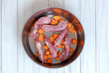 Top view on metal pet bowl with chicken necks and cut carrot inside. Beginning of natural organic daily meal ration preparation, BARF diet for dogs concept. Copy space, white wooden table background.