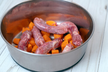 Metal pet bowl with chicken necks and cut carrot inside. Beginning of natural organic daily meal ration preparation, BARF diet for dogs concept. Copy space, white wooden table background.