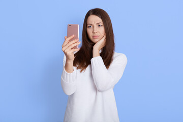 Fototapeta premium Confused young beautiful woman posing isolated over blue background, using mobile phone, having video call, hears unpleasant news, being upset, wearing white sweater.