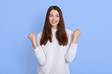 Fototapeta na wymiar Successful dark haired female, looks happy, clenches fists, accomplishes goal, exclaims finally victory, stands amused over blue background, excited lady dresses white sweater.